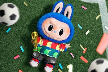 Mascote azul sorridente, vestindo uniforme esportivo colorido, segurando a Copa do Mundo de Ouro no campo de futebol com bola e confete