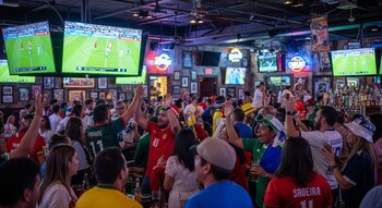 Bar de Dallas lleno de personas animando y levantando los brazos, viendo un partido de fútbol en grandes pantallas. Fondo con barra de licores y botellas.