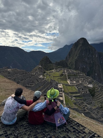 Tres personas sentadas de espaldas sobre una rejilla de metal observan un vasto complejo arqueológico de piedra y montañas bajo un cielo gris