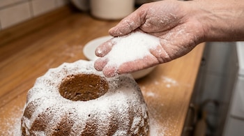 Primer plano de una mano espolvoreando azúcar glasé sobre una torta Bundt marrón oscura con forma de anillo, sobre una superficie de madera.