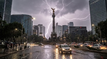 Avenida urbana de noche bajo la lluvia con relámpagos en el cielo, carros circulando y peatones con paraguas frente al Ángel de la Independencia y edificios.