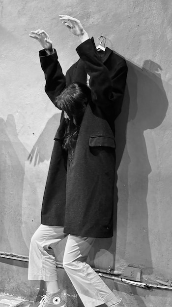 Fotografía en blanco y negro de una persona agachada con cabello largo, parcialmente cubierta por un abrigo oscuro colgado, contra una pared texturizada