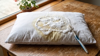 Una almohada blanca sobre una mesa de madera tiene una mancha amarilla cubierta de pasta blanca espumosa junto a un cepillo de dientes.