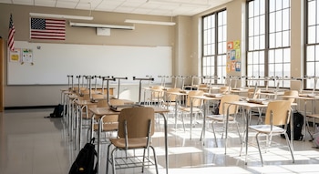 Aula moderna con escritorios y sillas de madera clara y metal organizados en filas, frente a un pizarrón blanco con una bandera de Estados Unidos y ventanas grandes.