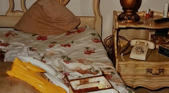 Dormitorio con cama, almohada, mesita de noche. Lámpara, teléfono fijo, reloj digital y esmaltes de uñas sobre la mesita. Bolsa amarilla y caja abierta en la cama