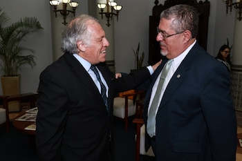 Luis Almagro, de traje negro, sonríe y toca el hombro de Bernardo Arévalo, de traje azul, en una sala con decoración clásica