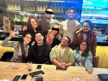 Un grupo de ocho personas, incluyendo Alberto "N" con el rostro censurado, posa sonriente alrededor de una mesa de madera en un restaurante con estanterías de botellas al fondo