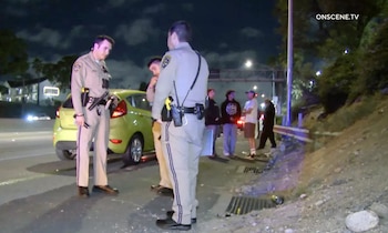 Dos agentes de policía en uniforme están junto a un coche verde claro en una carretera de noche. Varias personas están de pie cerca, con tráfico y vegetación a la derecha