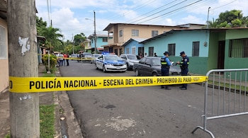 Cinta amarilla con la leyenda escena del crimen, una valla metálica, dos policías con libretas y una patrulla en una calle.