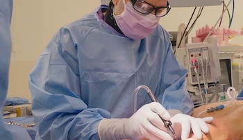 Un cirujano con bata azul, mascarilla, gafas protectoras y guantes sostiene un instrumento quirúrgico durante una cirugía