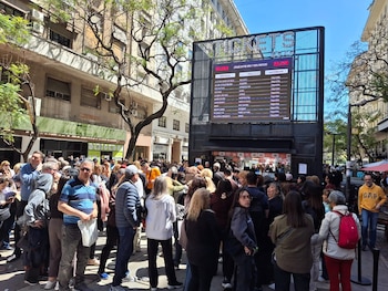 Un grupo numeroso de personas forma una fila frente a una estructura negra con pantalla gigante que promociona funciones teatrales en una calle