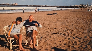 Dos hombres sentados en sillas sobre la arena de una playa. Al fondo se ven el mar con olas, un muelle y edificaciones costeras