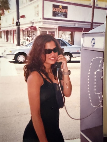 Una mujer con gafas oscuras y vestido negro habla por un teléfono público en una acera, con comercios y un vehículo al fondo