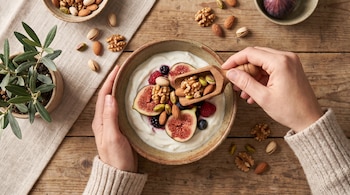 Vista superior de unas manos que sirven nueces y almendras en un tazón con yogur e higos sobre una mesa de madera.