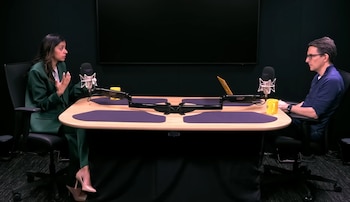 La Dra. Trisha Pasricha y un hombre sentados en una mesa de estudio de podcast con dos micrófonos, tazas amarillas y un portátil, sobre un fondo oscuro