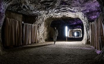 Un hombre con un mono blanco camina por un pasillo rocoso dentro de una cueva oscura con cortinas laterales, iluminación vertical brillante y techos rocosos