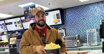Hombre barbudo con gorra de béisbol de los Yankees y chaqueta marrón sonríe mientras sostiene un gran cubo de palomitas en un puesto de comida