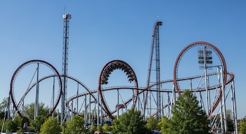 Montaña rusa Intimidator 305 con vías rojas, soportes blancos, un bucle vertical y torres altas, bajo un cielo azul claro, con árboles verdes en la base.