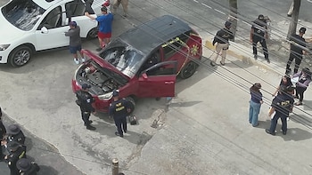 Un vehículo hatchback rojo con capó y puertas abiertas y cinta policial amarilla, un SUV blanco, agentes de policía y personas en una calle
