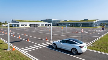 Pista de pruebas de manejo con conos naranjas, señales de tráfico y marcas viales; un coche blanco estacionado en la línea de salida y edificios modernos de techos verdes.