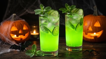 Dos vasos de bebida verde con hielo, pepino y menta junto a calabazas de Halloween iluminadas sobre una mesa de madera.