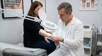Un dermatólogo vestido de blanco examina el antebrazo de una paciente con una lupa iluminada en una consulta médica, con un póster de fondo.