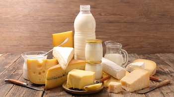 Una gran variedad de productos lácteos como leche en botella, quesos de diferentes formas y colores, yogures en vasos y mantequilla, sobre una mesa de madera.