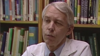 Un hombre de mediana edad con cabello gris y bata de laboratorio mira hacia la izquierda, con un fondo de estanterías llenas de libros