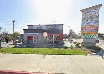 Exterior de un Carl's Jr con fachada gris y roja, frente a un poste alto con letreros de varios negocios, y una calle con un coche a la derecha
