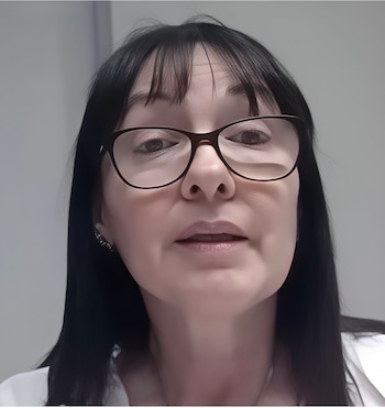Primer plano de una mujer con cabello negro liso y flequillo, usando gafas de montura oscura, mirando al frente. Ojos marrones, labios rosados y blusa blanca visible