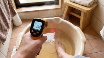 Una mano sostiene un termómetro digital que apunta a un recipiente con agua humeante y un pie dentro. El termómetro marca 36.0 grados Celsius.