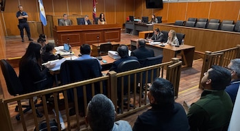 Tres jueces sentados en un estrado, abogados frente a escritorios con expedientes, un policía de pie y personas en la parte posterior