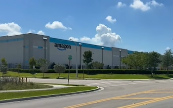 Amplio edificio de Amazon gris y azul con su logo en la fachada, visto desde una carretera con césped y árboles verdes bajo un cielo azul con nubes