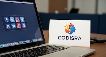 Una computadora portátil abierta sobre una mesa con íconos de redes sociales en la pantalla y una tarjeta con el logo de CODISRA al lado.