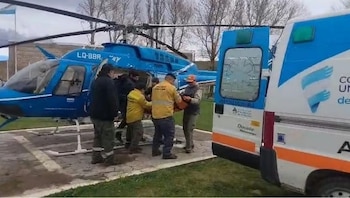 Varias personas trasladan una camilla entre un helicóptero azul y una ambulancia estacionados en un helipuerto