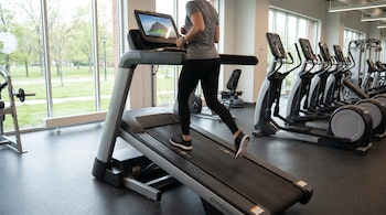 Una mujer corre en una cinta inclinada en un gimnasio. Se ven grandes ventanas con árboles verdes afuera y otras máquinas de ejercicio.