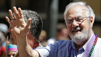 Barney Frank, hombre de barba y cabello blanco, gafas y camisa azul a cuadros, sonríe y saluda con la mano. Lleva collares de colores