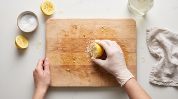 Manos sobre una tabla de cortar de madera con medio limón y sal, junto a un cuenco de sal, limones cortados y un paño
