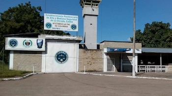 Fachada y portón de un centro penitenciario con una torre de vigilancia, muros de piedra y letreros institucionales bajo un cielo despejado