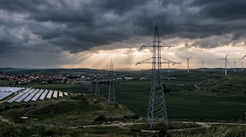 Cielo nublado con rayos de sol. Campos verdes, torres de alta tensión con cables, paneles solares. Molinos de viento en las colinas y un pueblo con iglesia al fondo.