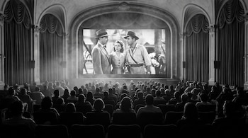 Auditorio de cine lleno, personas sentadas de espalda, pantalla con película en blanco y negro, telones laterales, columnas decoradas, haz de luz.