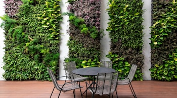 Terraza con paredes cubiertas de jardines verticales de follaje verde y morado, una mesa redonda oscura y cuatro sillas con asientos tejidos en un suelo de madera.