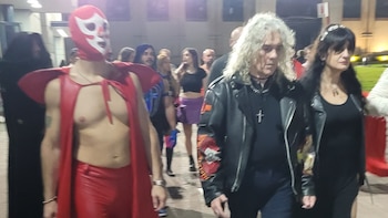 Hombre enmascarado con capa roja de luchador y otro hombre de cabello rizado blanco con chaqueta de cuero. Fondo borroso con personas en un evento interior