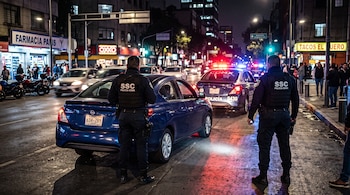 Dos agentes de policía con chalecos negros junto a un automóvil azul detenido en una calle nocturna ante una patrulla con luces encendidas.