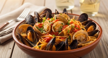 Un plato de espagueti con mejillones y almejas, bañado en salsa de tomate y espolvoreado con perejil, se ve sobre una mesa de madera con utensilios.