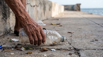 Mano con arrugas que toma una botella de plástico transparente del suelo agrietado, con un muro de hormigón y el mar de fondo.