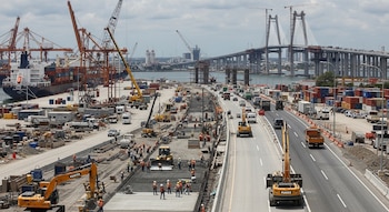Vista aérea de un sitio de construcción con maquinaria pesada, trabajadores, una autopista con vehículos y un puente atirantado sobre el agua en Veracruz.