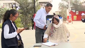 Una mujer en silla de ruedas con un poncho claro y un pañuelo estampado es asistida por un hombre, mientras otra mujer mira su teléfono en un centro de votación