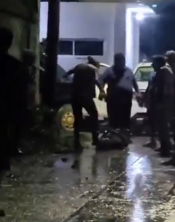 Varios hombres en una calle mojada de noche junto a una persona tendida en el suelo y dos vehículos estacionados