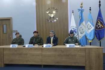 Cuatro personas sentadas tras una mesa con letreros de identificación, frente a una pared con el escudo de la Fuerza Aérea Guatemalteca